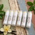 Six Wild Herb lip balm tubes arranged on a rustic painted wood surface with green leaves and a vanilla flower around them. The labels show different scents: Arctic Orange, Bubble Gum, Cotton Candy, Peppermint, Wild Water, and French Vanilla.
